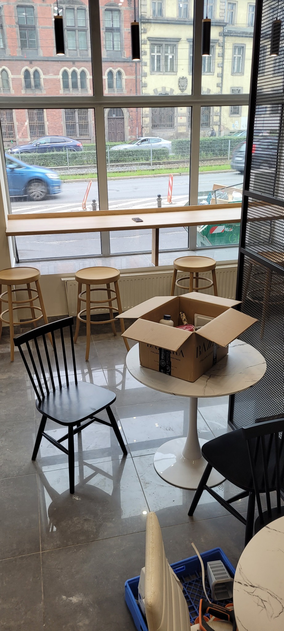 Wnętrze lokalu z dużym oknem, widok na ulicę, stoliki z krzesłami i karton z materiałami do wykończenia wnętrz, prawdopodobnie farby i akcesoria malarskie.