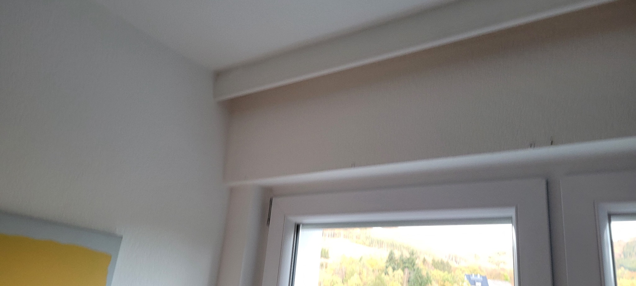 Fragment wnętrza: pomalowana na biało ściana z widocznym oknem, fragment sufitu z dekoracyjną listwą i żółty fragment ściany w rogu.