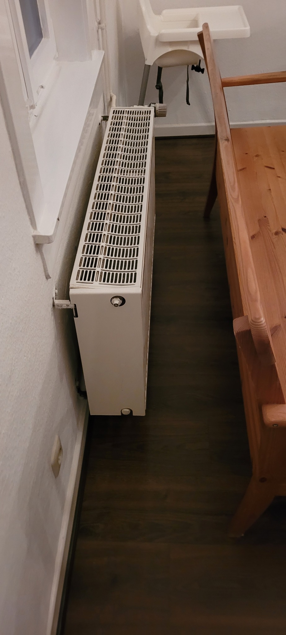 Biały, żebrowany grzejnik przymocowany do ściany obok okna, widoczna podłoga z ciemnego drewna i fragment drewnianego krzesła dziecięcego.