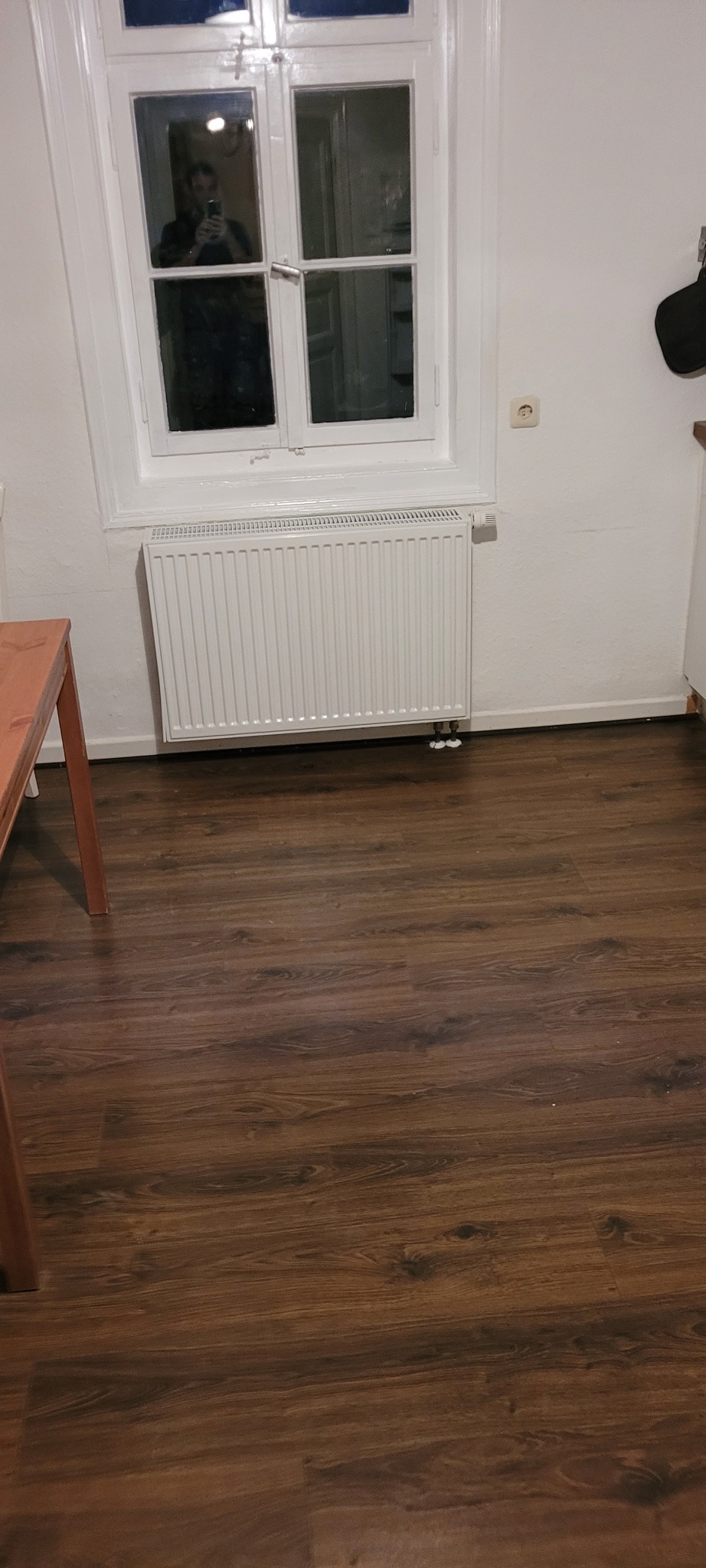 Wnętrze pokoju z białymi ścianami, drewnianą podłogą, grzejnikiem pod oknem i fragmentem stołu po lewej stronie. W oknie widoczne odbicie osoby robiącej zdjęcie.