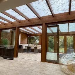 Drewniana konstrukcja z przezroczystym zadaszeniem, tworząca oszkloną przestrzeń z widokiem na ogród, wyposażona w meble wypoczynkowe.