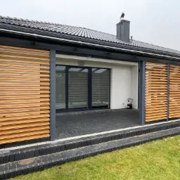 Nowoczesny taras z przesuwnymi, drewnianymi panelami żaluzjowymi w kolorze jasnego drewna, zamontowanymi na metalowej konstrukcji w kolorze antracytowym, prowadzącymi do przeszklonego wejścia...