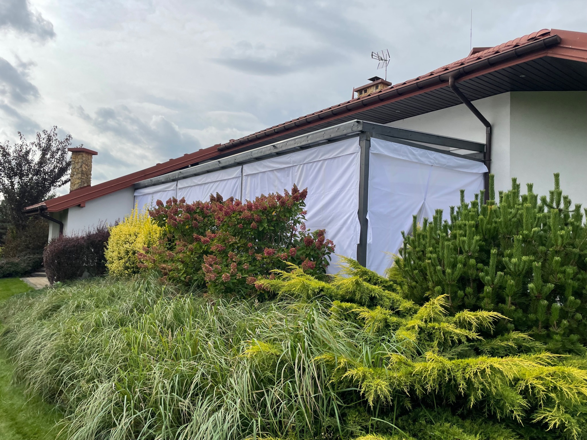Szara pergola z białymi zasłonami zamontowana przy domu, otoczona ozdobnymi krzewami i trawami, widok na dach i komin.