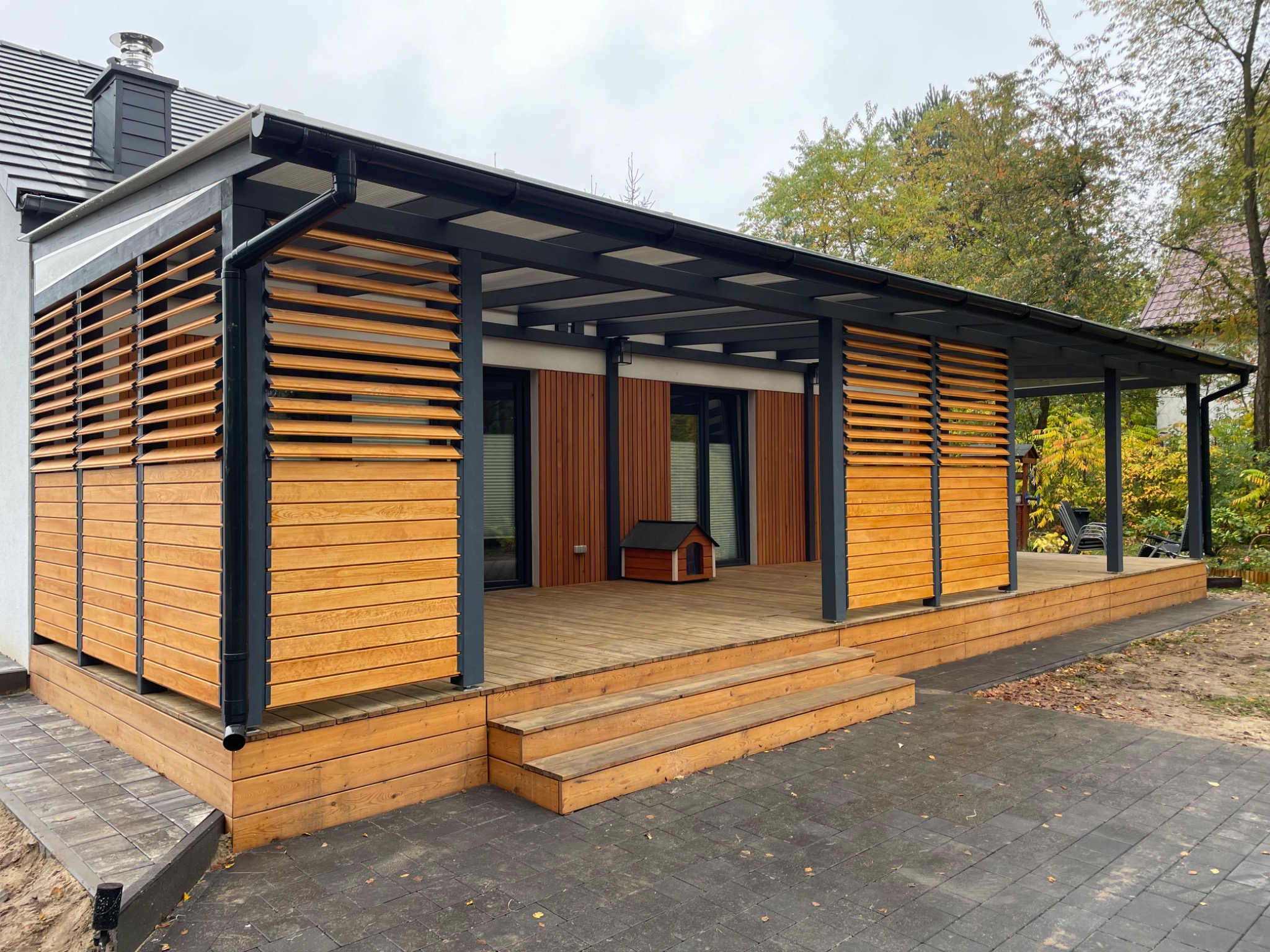 Nowoczesna konstrukcja z drewnianymi żaluzjami i metalowym szkieletem, z niskim dachem, drewnianym tarasem i schodami, z małą budą dla psa w tle.