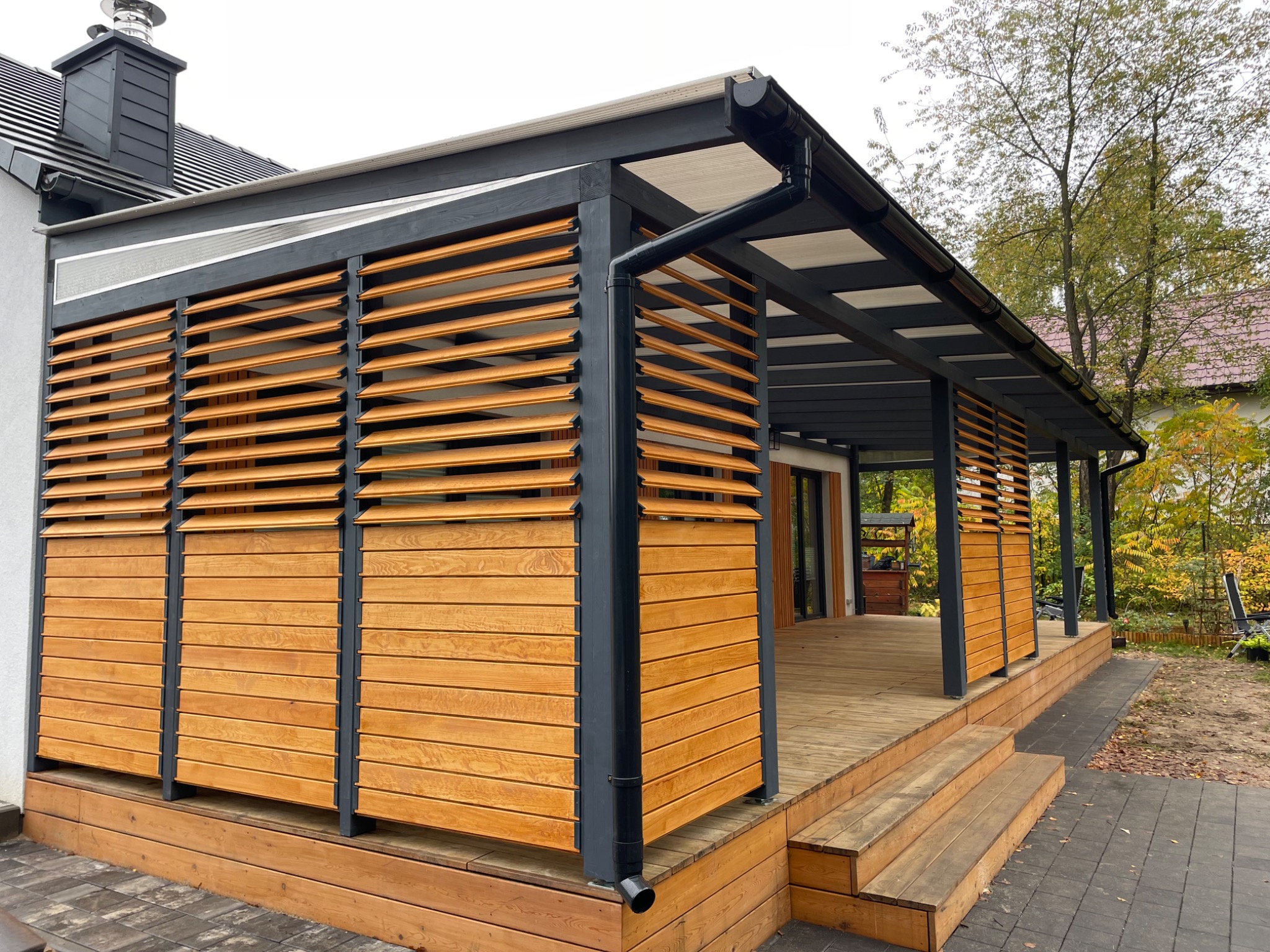 Zadaszona konstrukcja z drewnianymi żaluzjami i czarnymi metalowymi słupkami, połączona z budynkiem mieszkalnym, na drewnianym tarasie z kilkoma schodami.