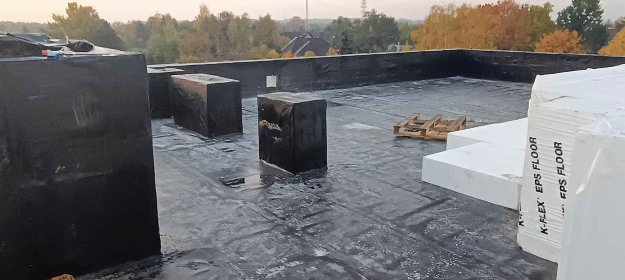 Płaski dach pokryty czarną membraną hydroizolacyjną, z zaizolowanymi kominami i zapasem styropianu EPS FLOOR, widok na jesienne drzewa w tle.
