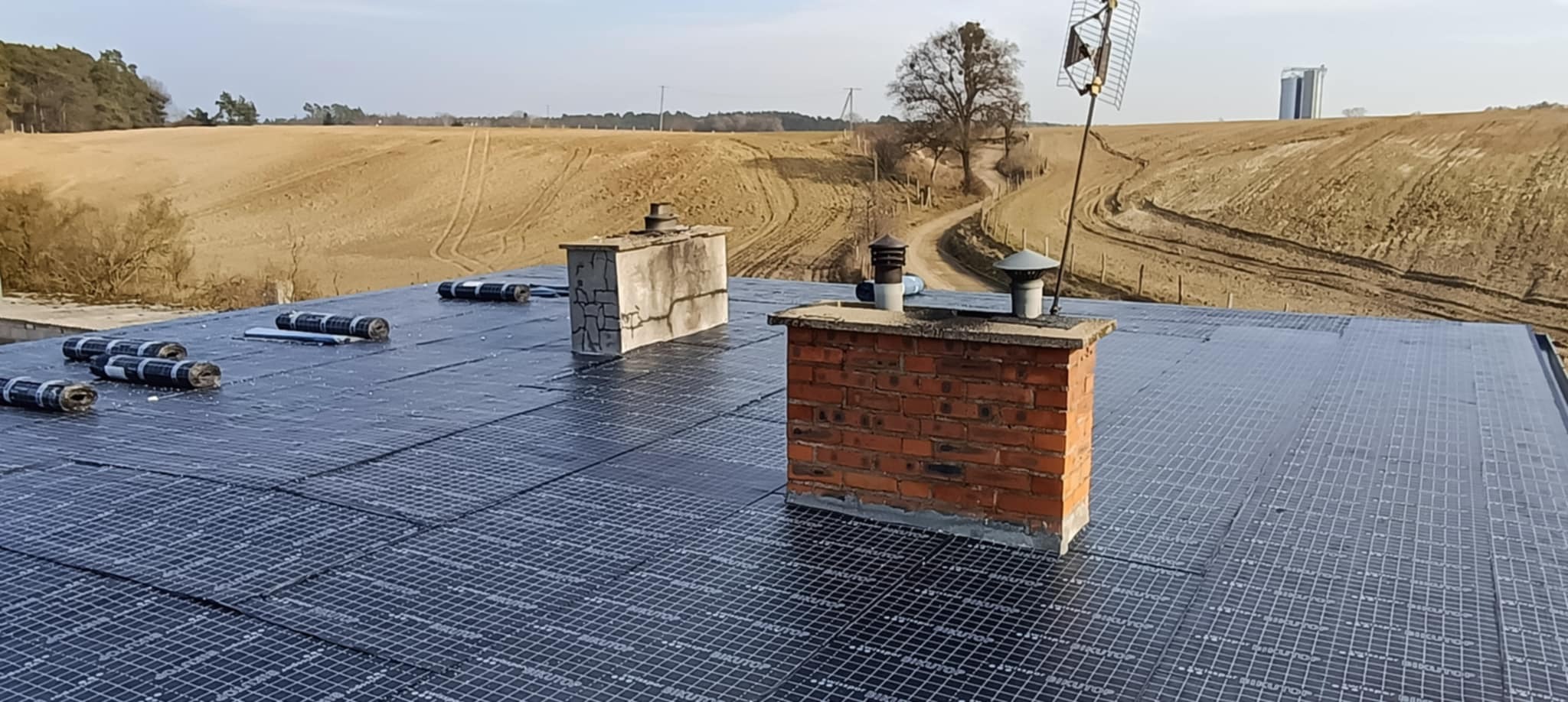 Widok z góry na dach pokryty membraną z widocznymi rolkami materiału, dwa kominy (jeden ceglany z anteną) i polami uprawnymi w tle.