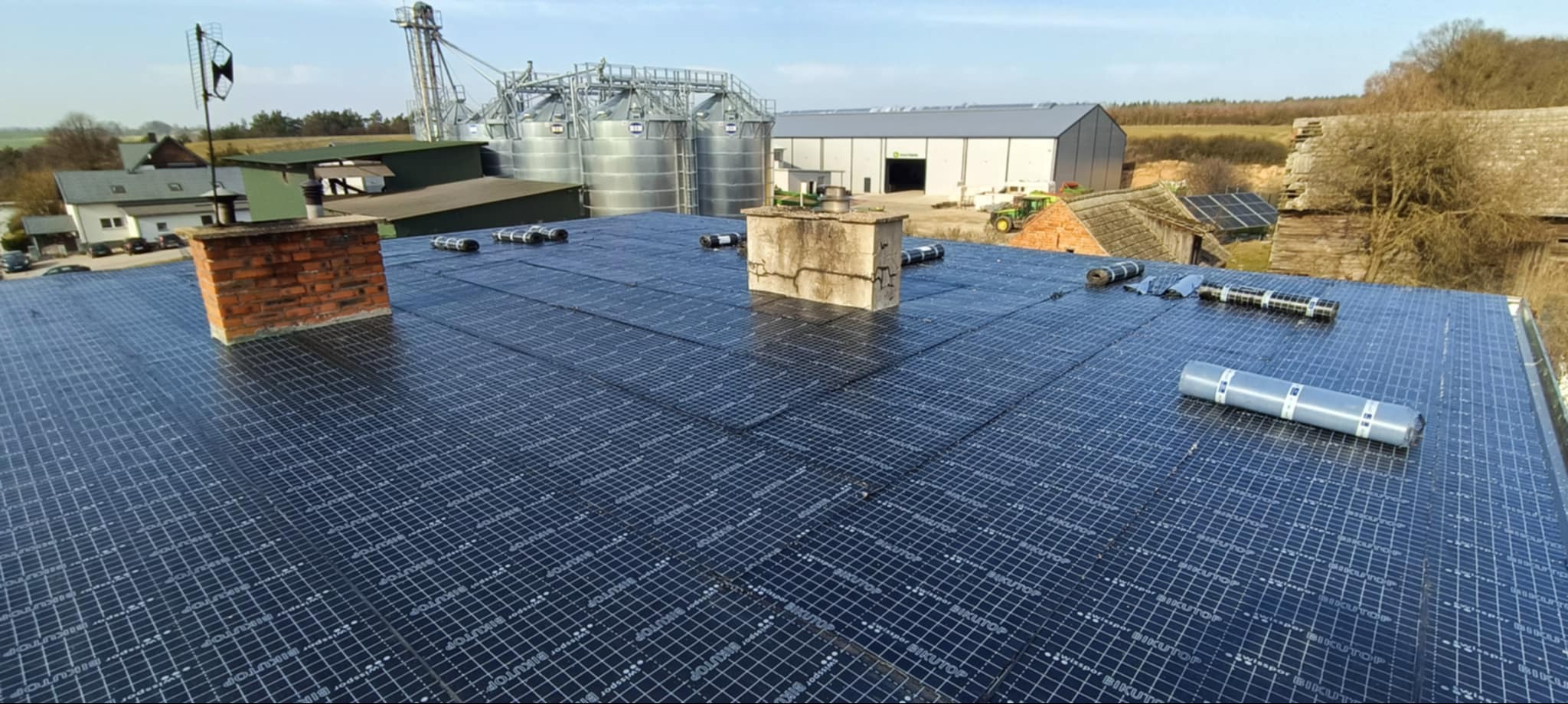 Nowe pokrycie dachu z papy bitumicznej z widocznymi kominami, rolkami papy i elementami wentylacji. W tle zabudowania gospodarcze i silosy.