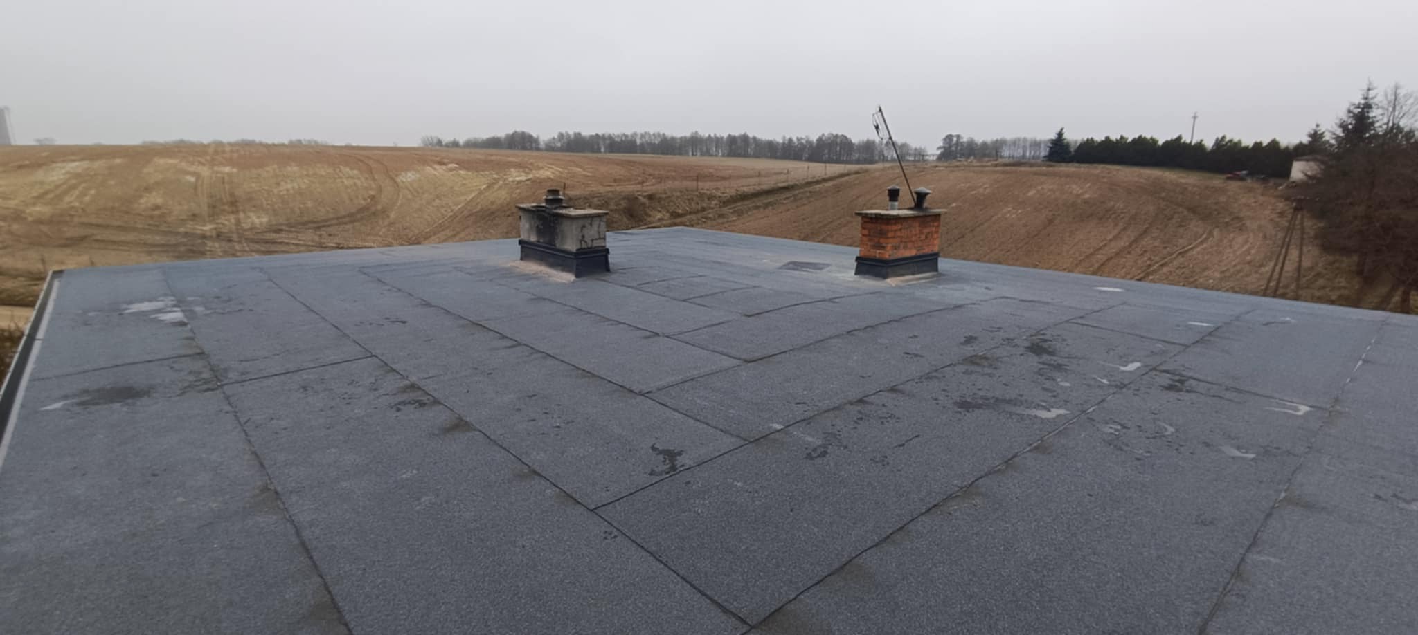 Widok z góry na płaski dach pokryty papą, z dwoma kominami - jeden murowany z cegły, drugi betonowy - na tle zaoranego pola i lasu w pochmurny dzień.