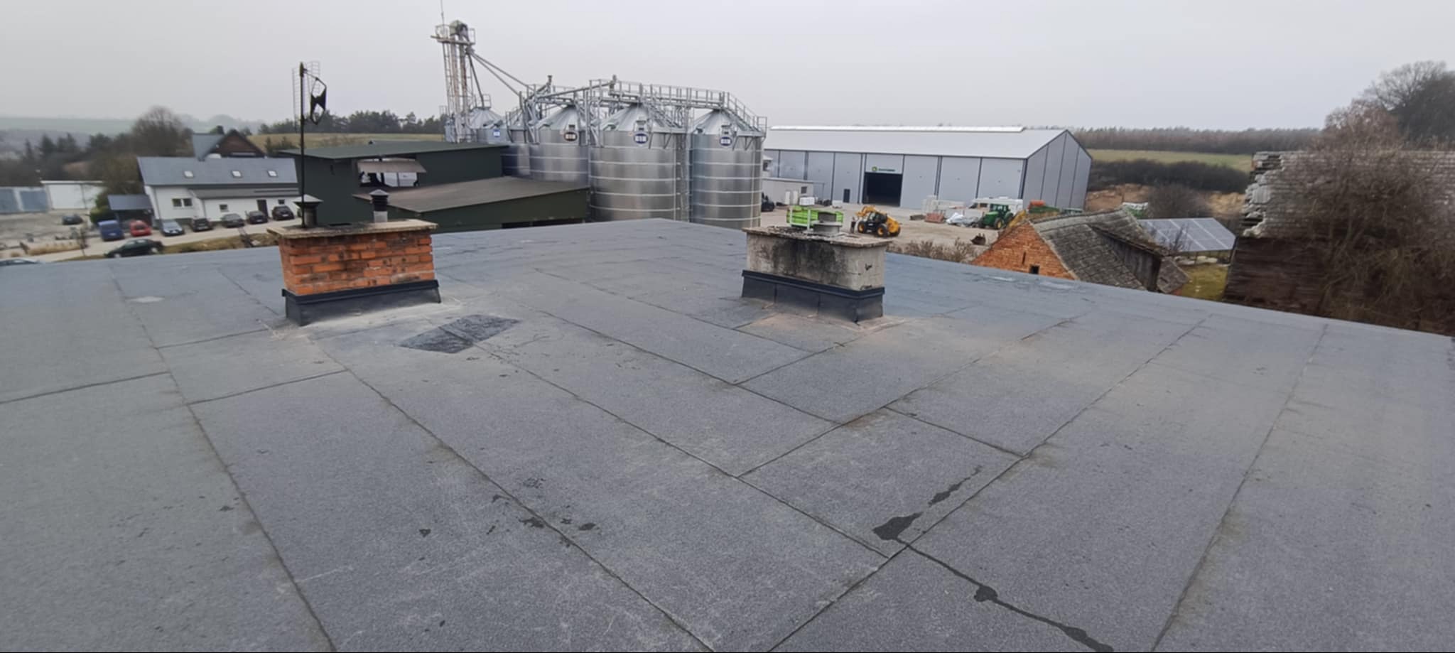 Widok z góry na pokryty papą dach z dwoma kominami, w tle zabudowania gospodarcze, silosy i hala.