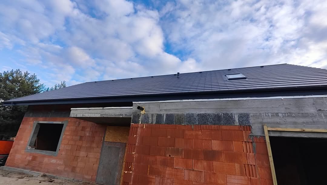 Nowy, grafitowy dach z oknem dachowym na budynku w trakcie budowy w Bydgoszczy, widok z dołu na tle pochmurnego nieba.