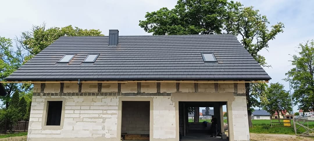 Nowy, grafitowy dach z dachówki ceramicznej z oknami połaciowymi i kominem na surowym budynku z białego bloczka, widoczne drewniane stemple podtrzymujące okap.