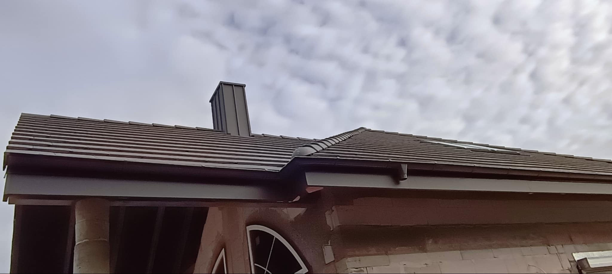 Brązowy dach z dachówki ceramicznej z ciemnymi obróbkami blacharskimi i rynnami, komin z blachy na tle pochmurnego nieba.