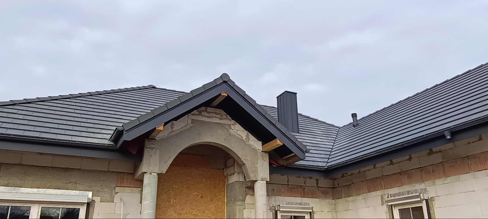 Nowy dach z ciemnej dachówki ceramicznej na domu w trakcie budowy, widoczne obróbki blacharskie i komin, surowy stan elewacji.