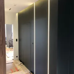 Korytarz w trakcie wykańczania, ściany pomalowane na ciemny kolor z wbudowanym oświetleniem LED, podłoga zabezpieczona kartonem, widoczna huśtawka w tle.