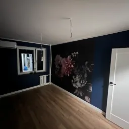 Pokój po remoncie: ciemnoniebieskie ściany, drewniana podłoga, fototapeta z motywem kwiatowym, białe drzwi i grzejnik, widoczne kable od oświetlenia sufitowego.