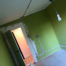 Leo Czerwiec - Pokój z zielonymi ścianami w trakcie remontu, widoczne kable elektryczne w bruzdach na ścianach, częściowo zakryte gipsem, oraz otwarta futryna drzwiowa z widokiem na jasne pomieszczenie.