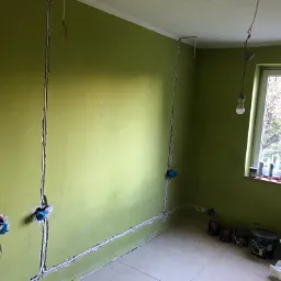 Zielony pokój w trakcie remontu: wycięte bruzdy w ścianach pod instalację elektryczną, puszki pod gniazdka, widoczne kable, farby i akcesoria malarskie przy oknie.