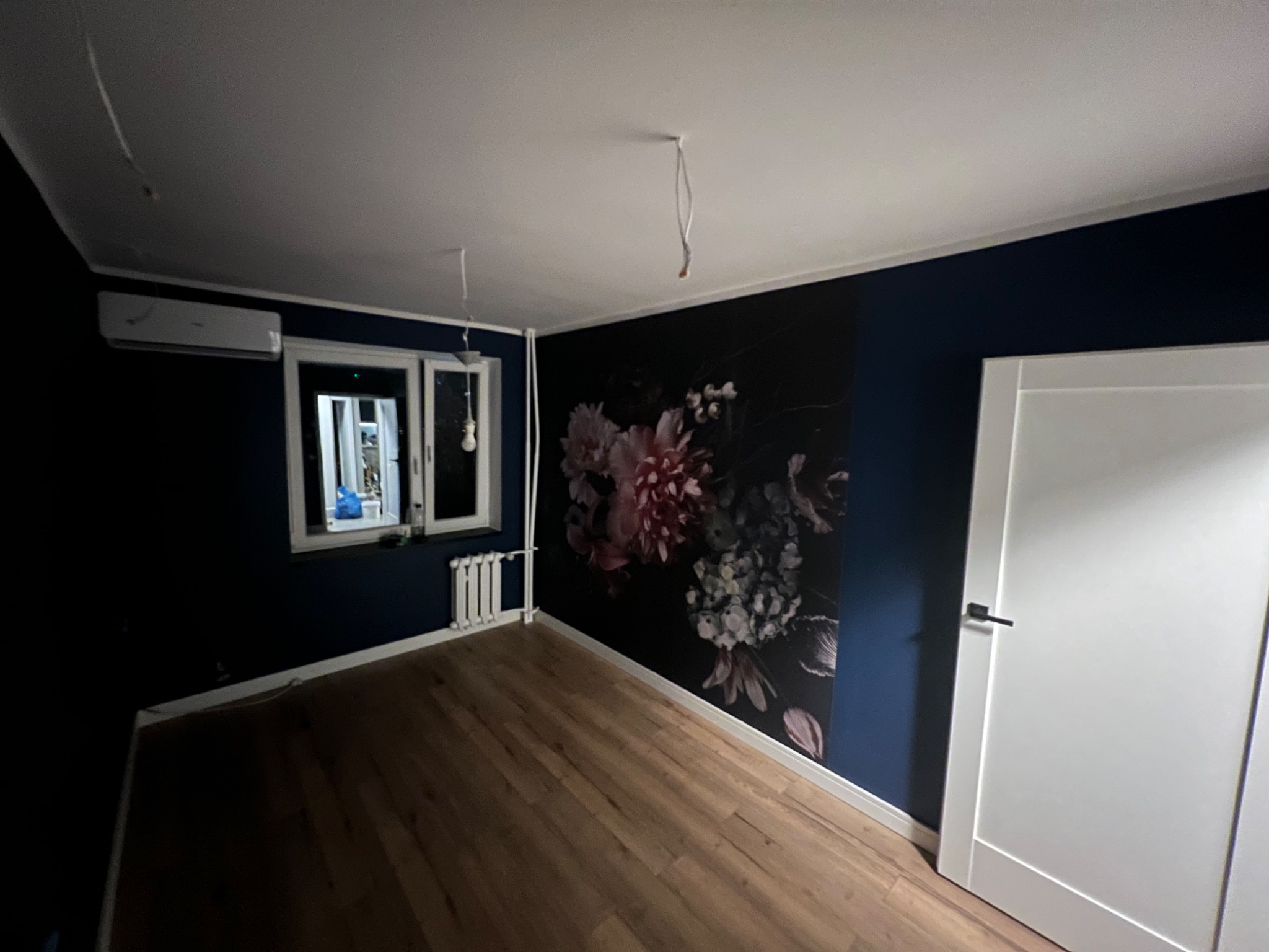 Pokój po remoncie: ciemnoniebieskie ściany, drewniana podłoga, fototapeta z motywem kwiatowym, białe drzwi i grzejnik, widoczne kable od oświetlenia sufitowego.