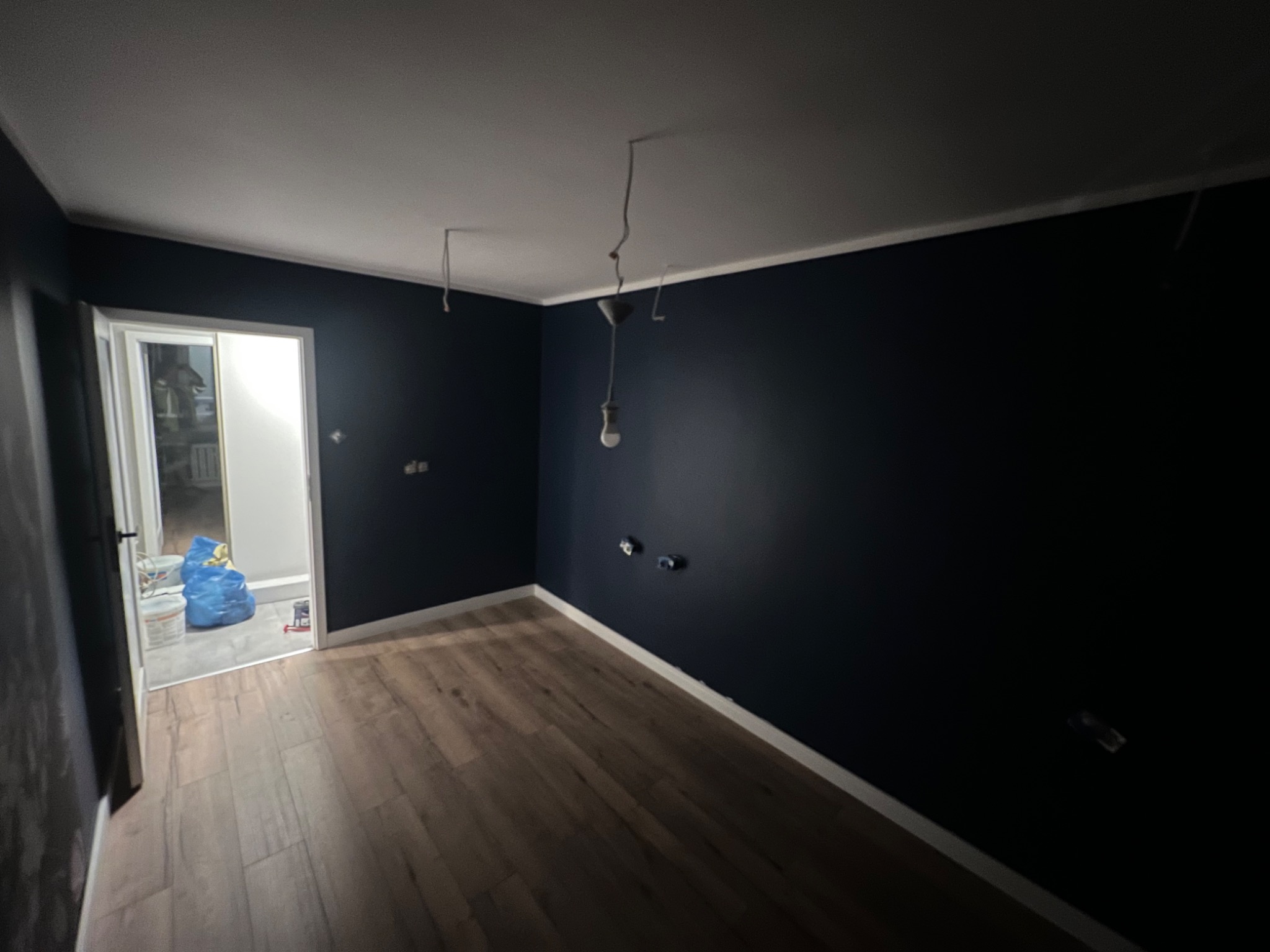 Pokój po remoncie z ciemnoniebieskimi ścianami, drewnianą podłogą, widoczne kable od oświetlenia zwisające z sufitu, w tle otwarte drzwi do kolejnego pomieszczenia z workami i wiadrami.