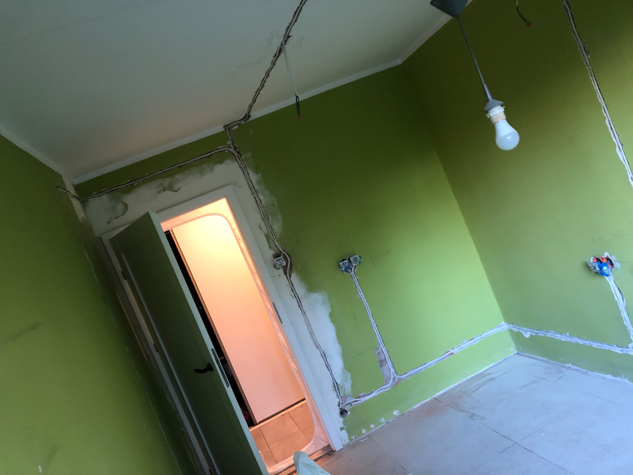 Pokój z zielonymi ścianami w trakcie remontu, widoczne kable elektryczne w bruzdach na ścianach, częściowo zakryte gipsem, oraz otwarta futryna drzwiowa z widokiem na jasne pomieszczenie.