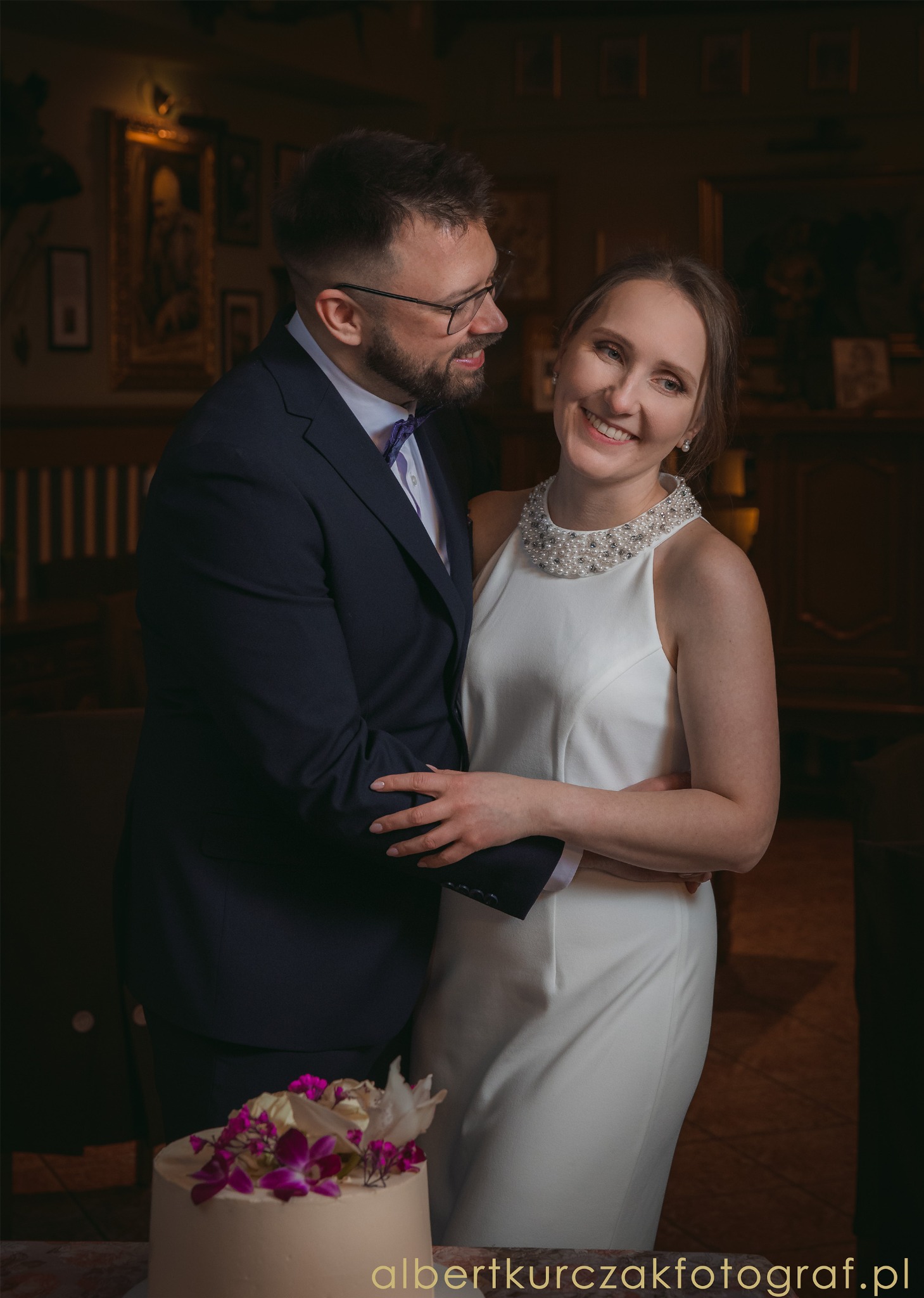 Uśmiechnięta para młoda w eleganckich strojach pozuje w dworskim wnętrzu z tortem weselnym. Pan w okularach i fioletowej muszce, pani w białej sukni z perłowym kołnierzem.