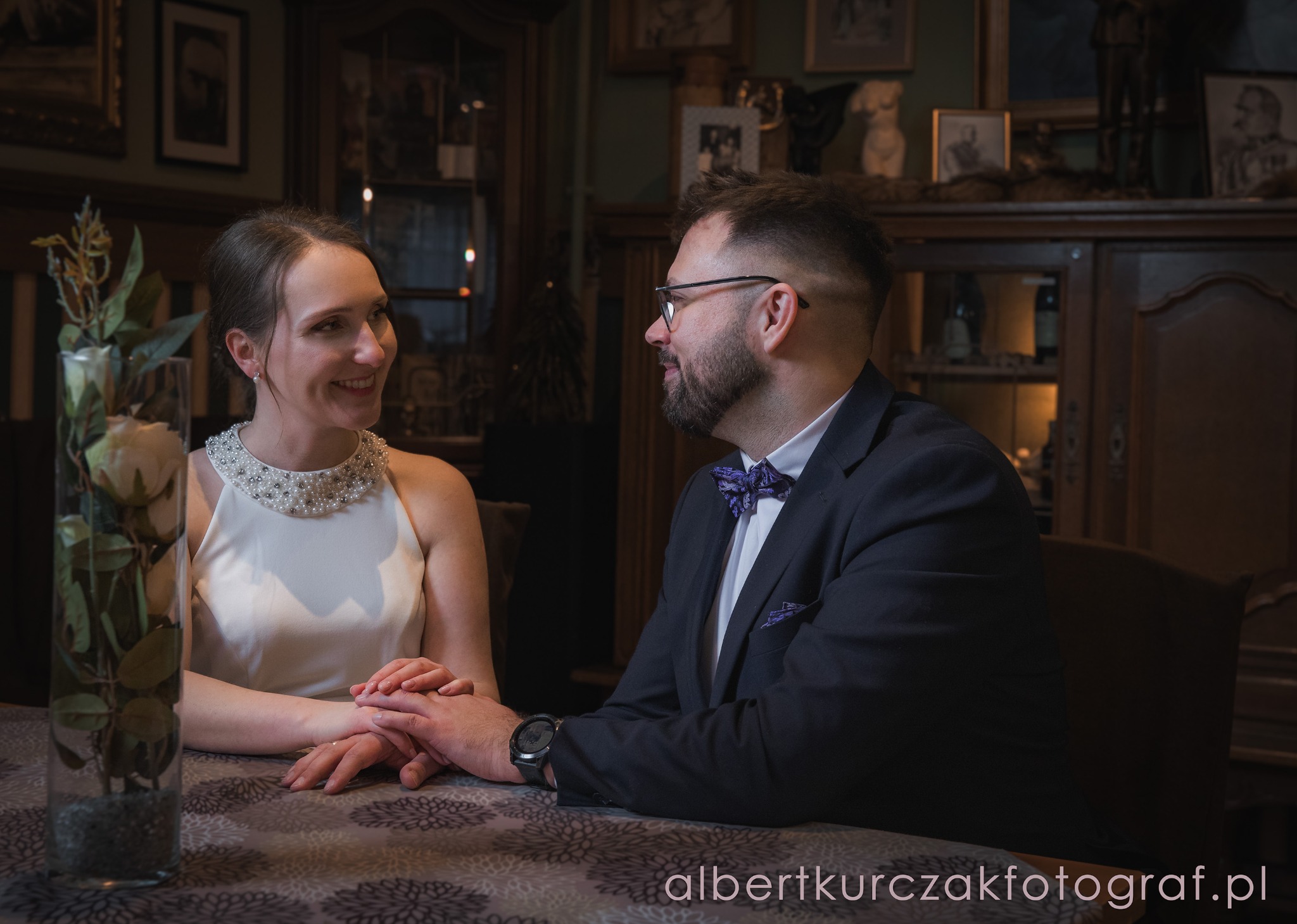 Uśmiechnięta para młoda siedzi przy stole w eleganckiej restauracji w stylu dworskim. Ona w białej sukni, on w ciemnym garniturze z muszką. Romantyczna atmosfera.