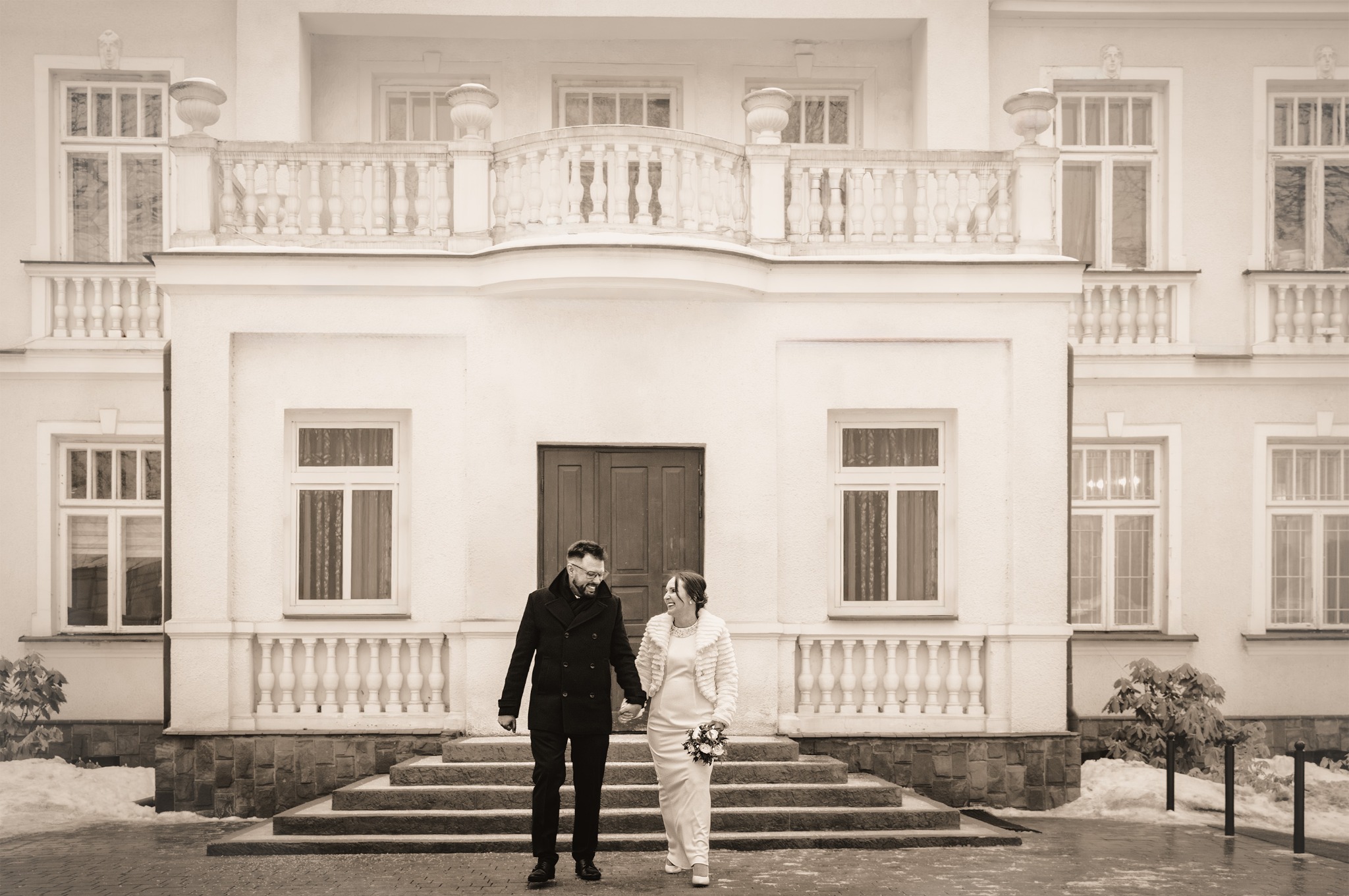 Para młoda, on w czarnym płaszczu, ona w białej sukni, trzymają się za ręce na tle klasycznego budynku z balkonem i kolumnami. Sepia, zimowa sceneria.