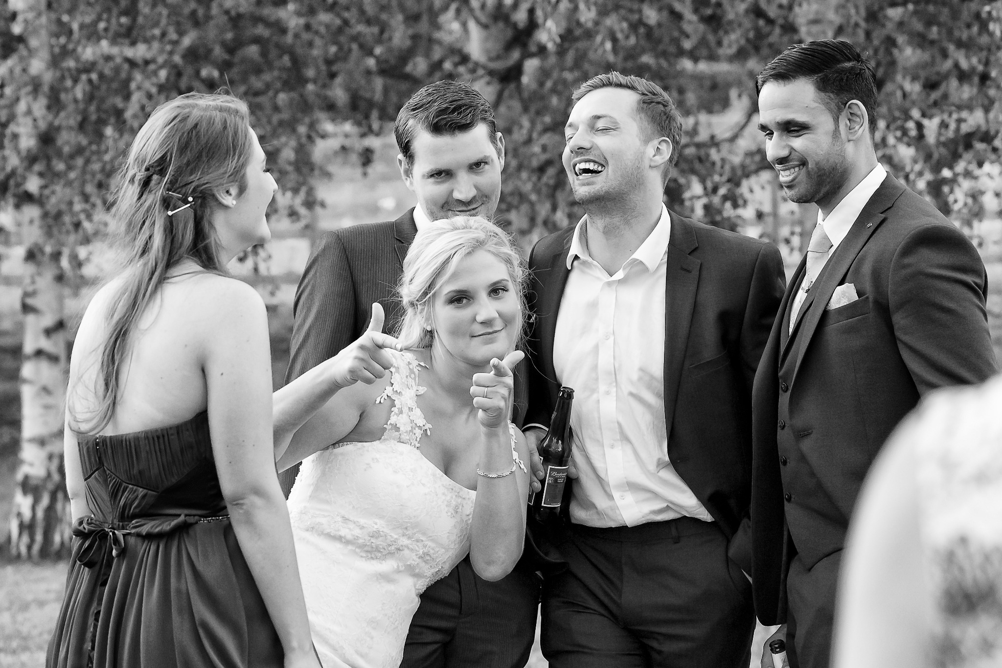 Czarno-białe zdjęcie grupowe z wesela: uśmiechnięta panna młoda z butelką piwa pozuje z gośćmi, wskazując palcami w stronę obiektywu.