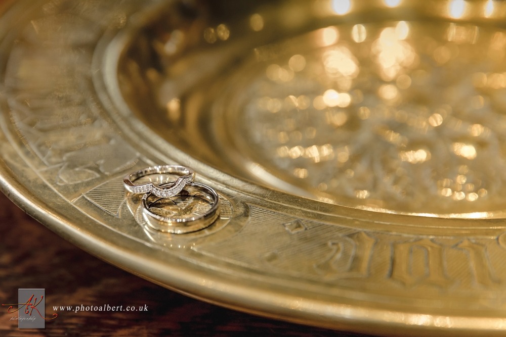 Dwa złote obrączki, jedna z diamentami, leżą na zdobionej, złotej tacy z wytłoczonymi napisami.