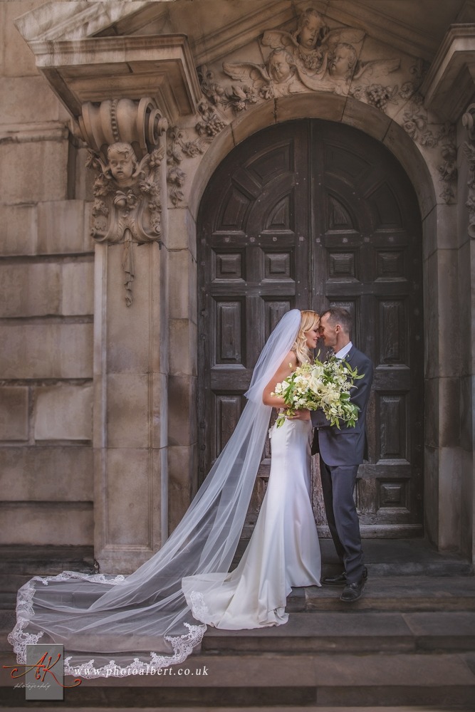 Para młoda stoi na schodach przed monumentalnymi, rzeźbionymi drzwiami, panna młoda w prostej sukni z długim welonem ozdobionym koronką, trzyma bukiet z białych kwiatów.