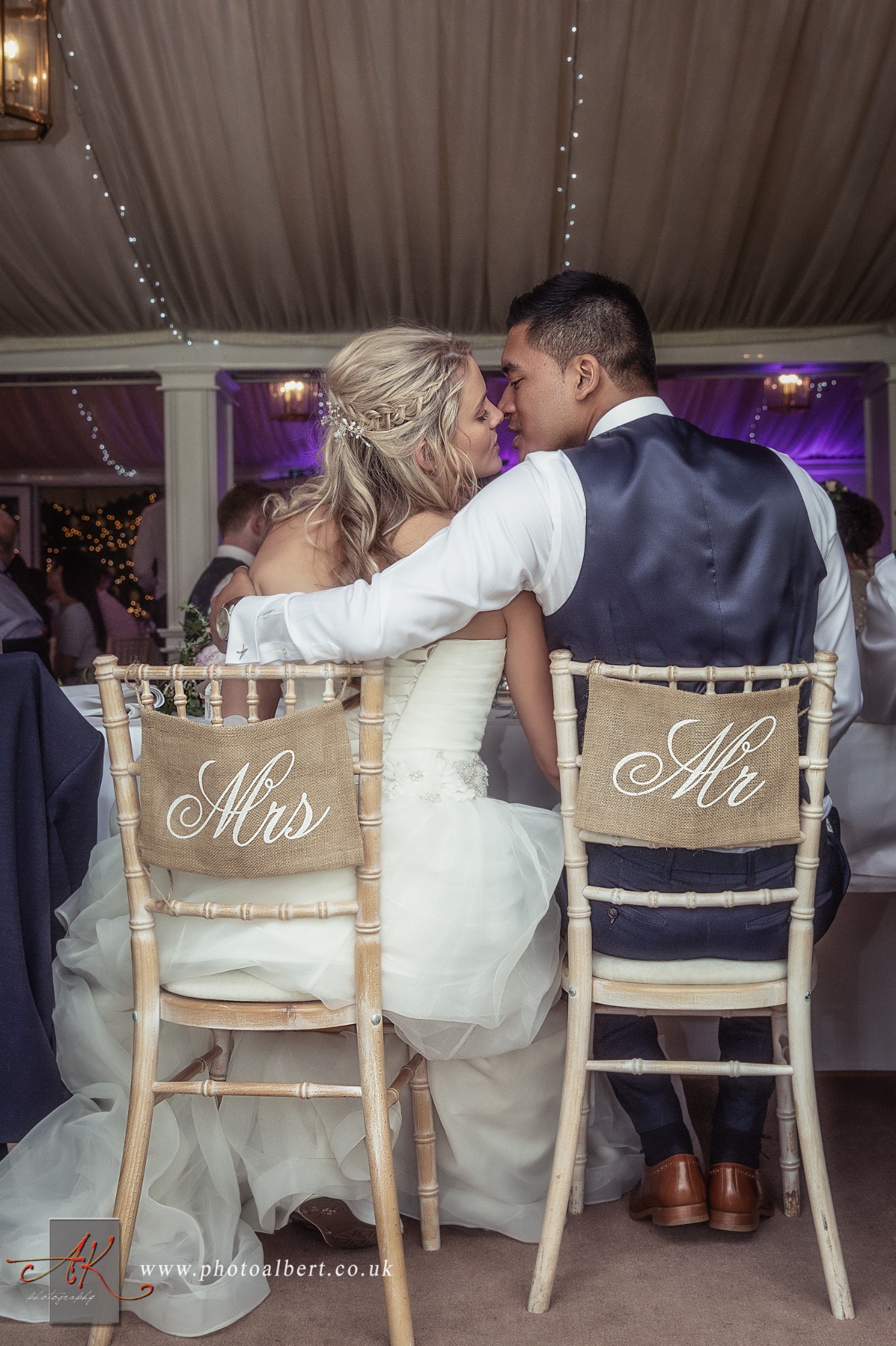 Panna młoda i pan młody w objęciach, siedzą na krzesłach z napisem 'Mrs' i 'Mr' podczas wesela w sali z dekoracjami świetlnymi.