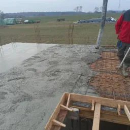 Wylewanie betonu na zbrojoną płytę fundamentową z widocznym deskowaniem otworu, w tle krajobraz wiejski z polami i zabudowaniami.