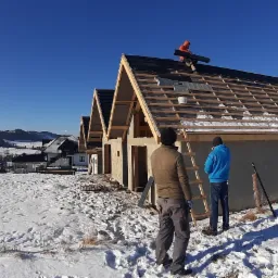 Wznoszenie szeregu domów jednorodzinnych w zimowym krajobrazie; widoczni dekarze układający pokrycie dachowe na jednym z budynków, z drewnianą konstrukcją więźby dachowej; ośnieżone pola...