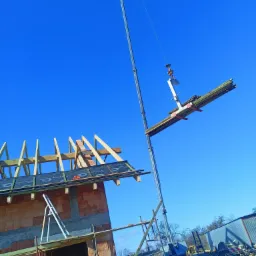 Budowa domu z cegły: dźwig podnosi drewniane elementy więźby dachowej na niebieskim tle nieba.