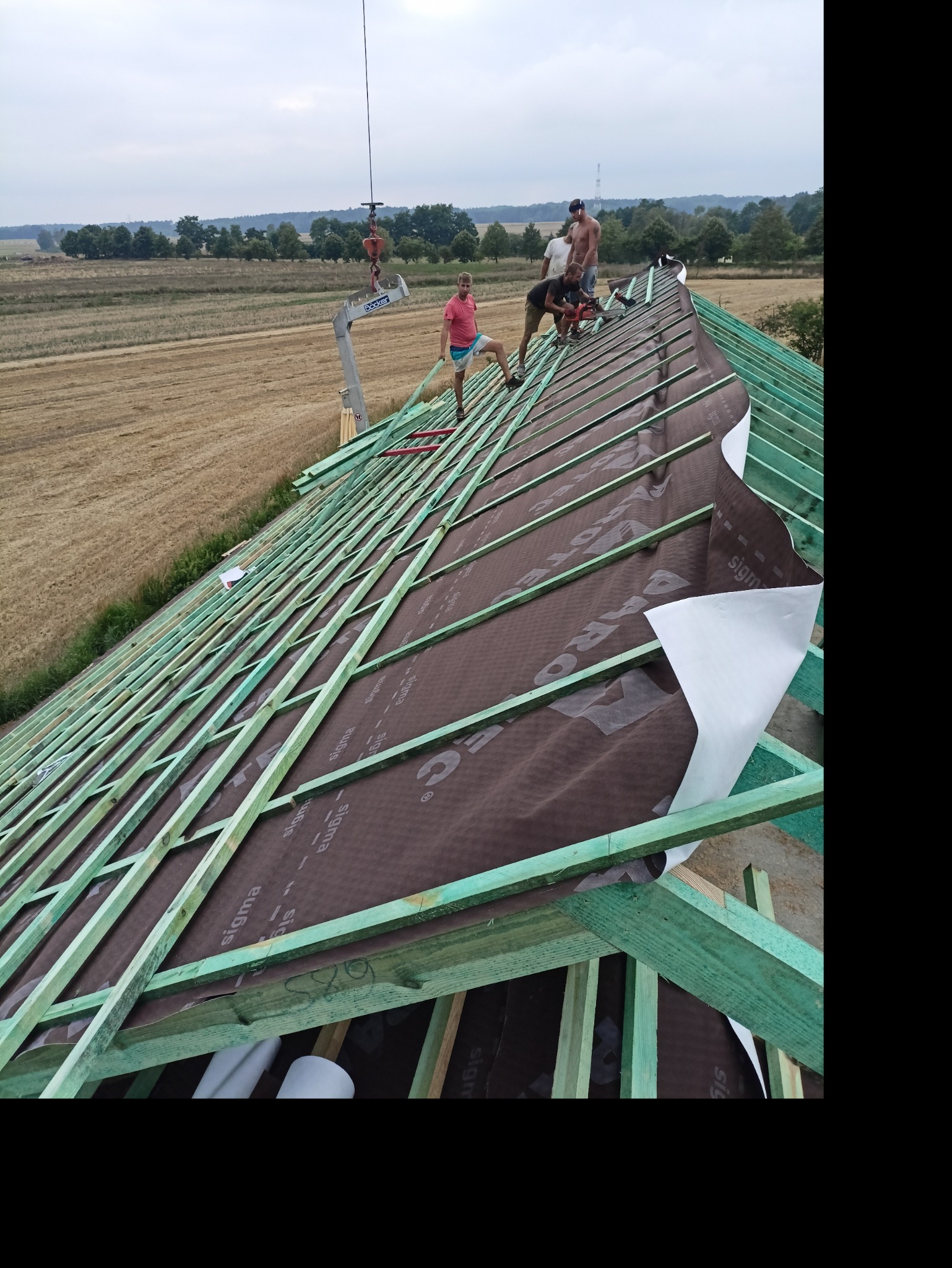 Montaż brązowej membrany dachowej na zielonej więźbie dachowej przez trzech mężczyzn, widok na polne tło.