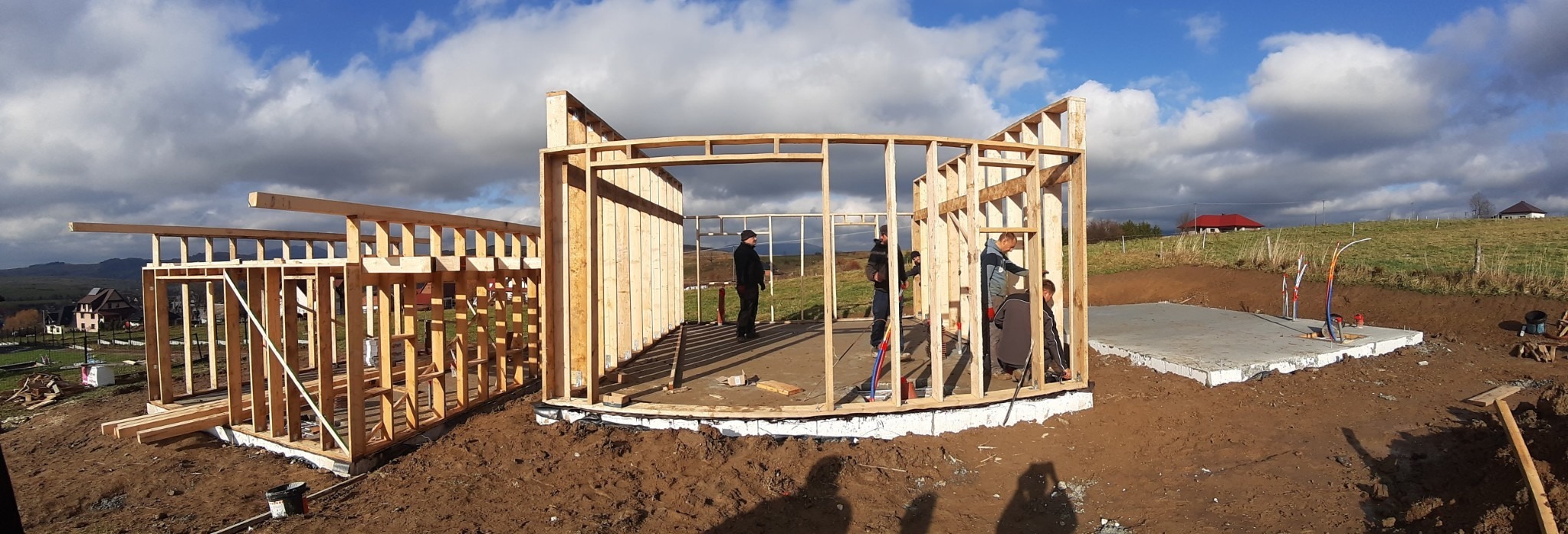Budowa domu szkieletowego w trakcie wznoszenia ścian, widoczna konstrukcja drewniana, fundamenty, pracownicy oraz malowniczy krajobraz w tle.