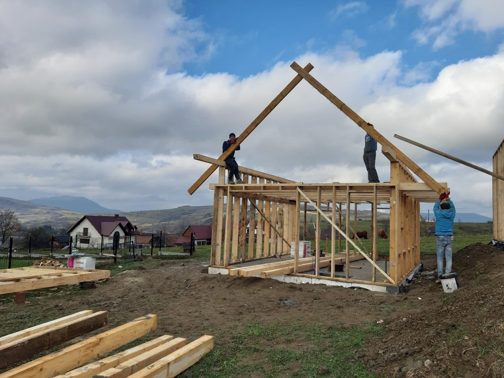 Trzech pracowników montuje drewnianą konstrukcję szkieletową domu na fundamencie w krajobrazie wiejskim, widoczna więźba dachowa w trakcie wznoszenia, w tle zabudowania i wzgórza pod pochmurnym...