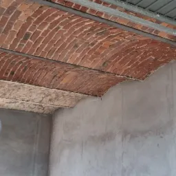 Pracownik w kombinezonie ochronnym i masce piaskuje łukowe sklepienie z cegły wewnątrz pomieszczenia, widoczne surowe ściany i instalacje.