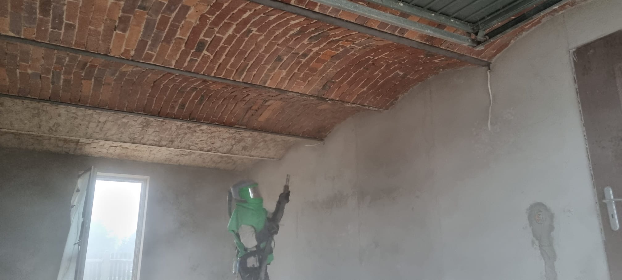 Pracownik w kombinezonie ochronnym podczas czyszczenia ceglanego sklepienia wewnątrz budynku za pomocą strumienia ściernego.