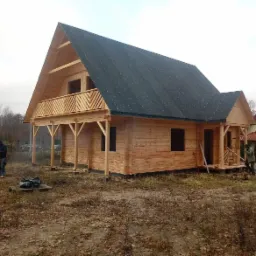 Surowy drewniany dom z ciemnym dachem w trakcie budowy, widoczni dwaj pracownicy na zewnątrz, niezagospodarowany teren wokół.