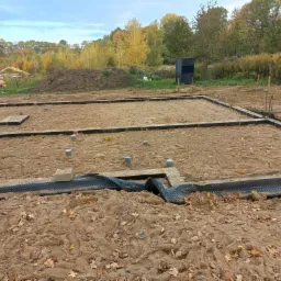 Przygotowany teren pod fundamenty domu z widoczną czarną folią hydroizolacyjną i wystającymi rurami kanalizacyjnymi, w tle jesienne drzewa.