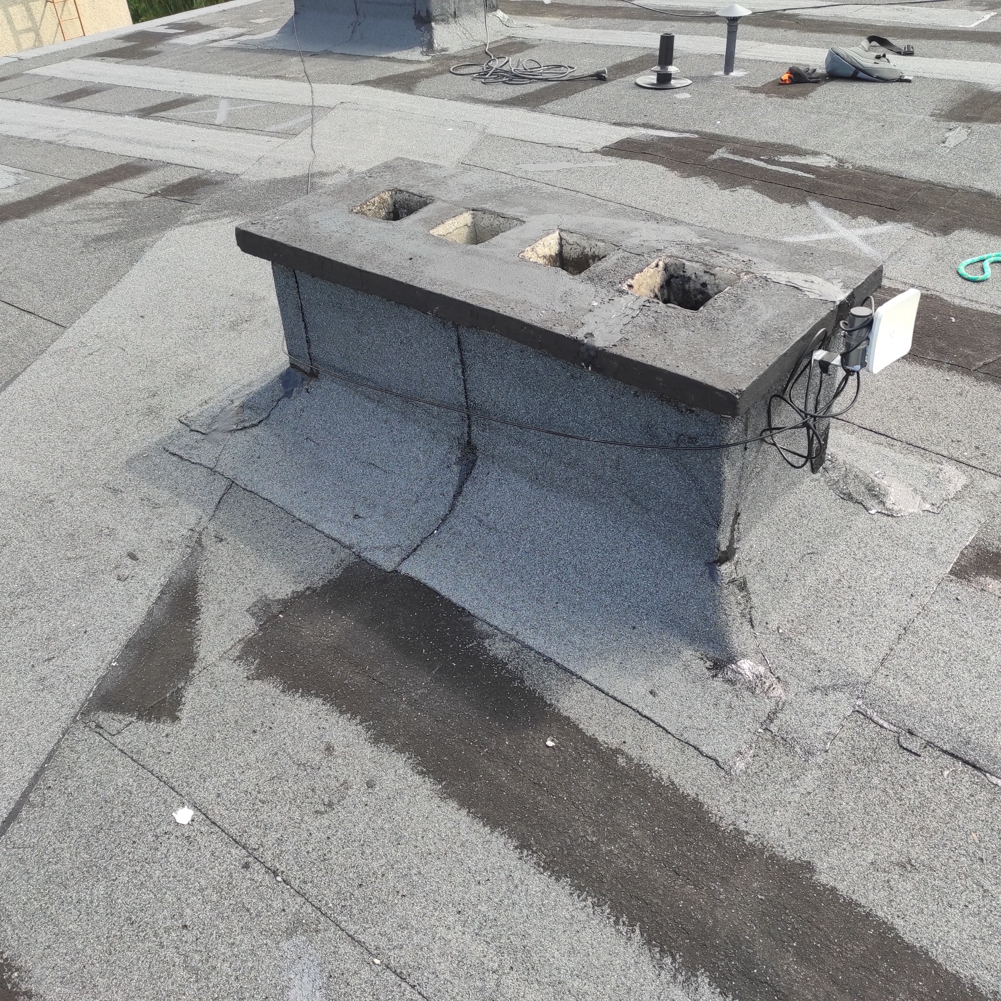 Płaski dach pokryty papą z kominem z trzema otworami, widoczne ślady napraw i akcesoria montażowe.