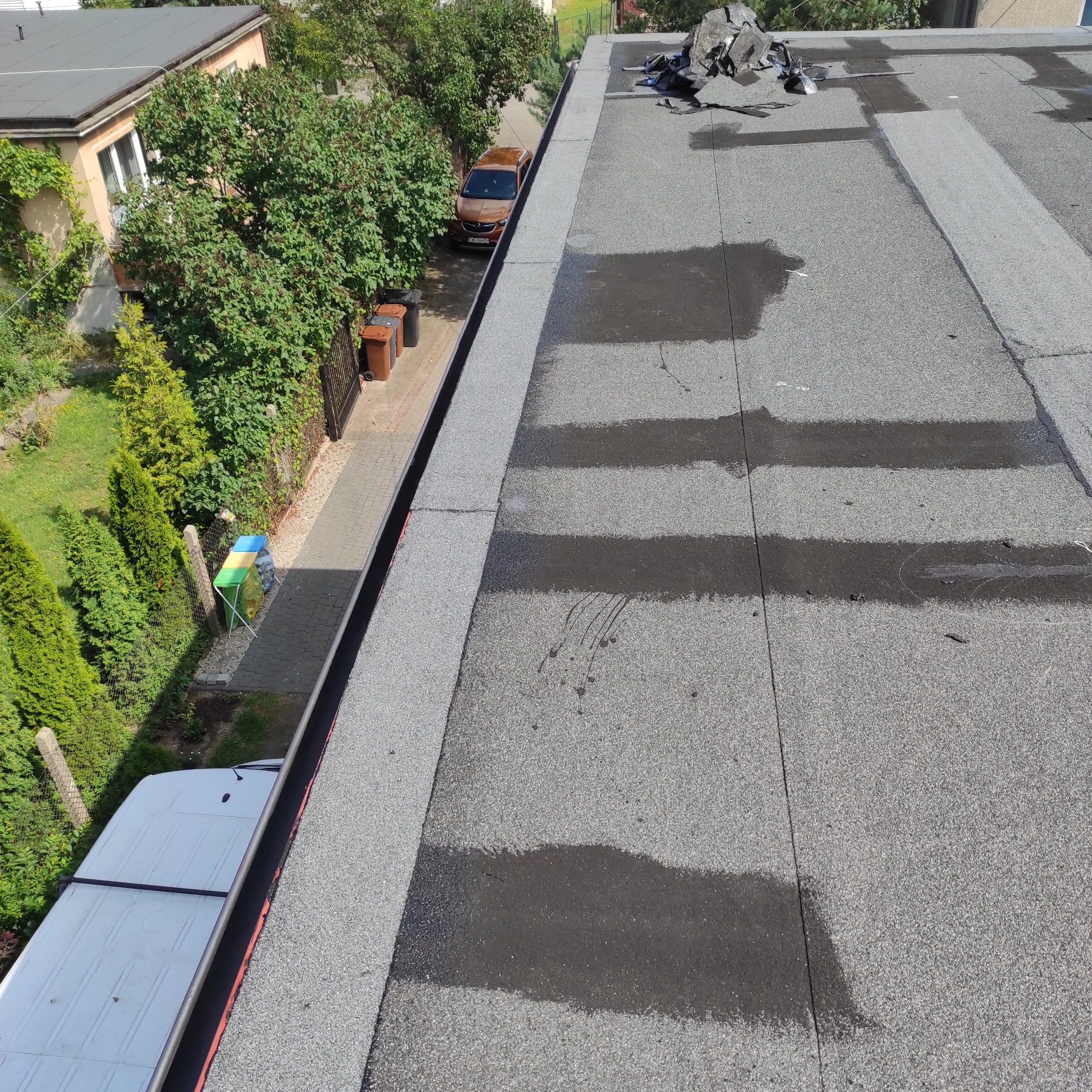 Widok z góry na płaski dach pokryty papą, z widocznymi śladami po wodzie, obok rynna i fragmenty starych materiałów dekarskich oraz otoczenie budynku z zielenią