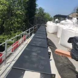 Czarna, tymczasowa ścieżka z płyt na dachu z widocznymi śladami butów, zabezpieczona barierkami, obok worki z materiałami budowlanymi i cegły.