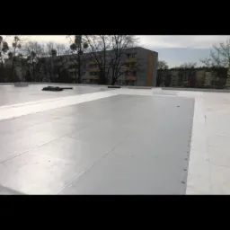 Szary, płaski dach z widocznymi łączeniami i elementami mocującymi, w tle budynek mieszkalny i drzewa bez liści.