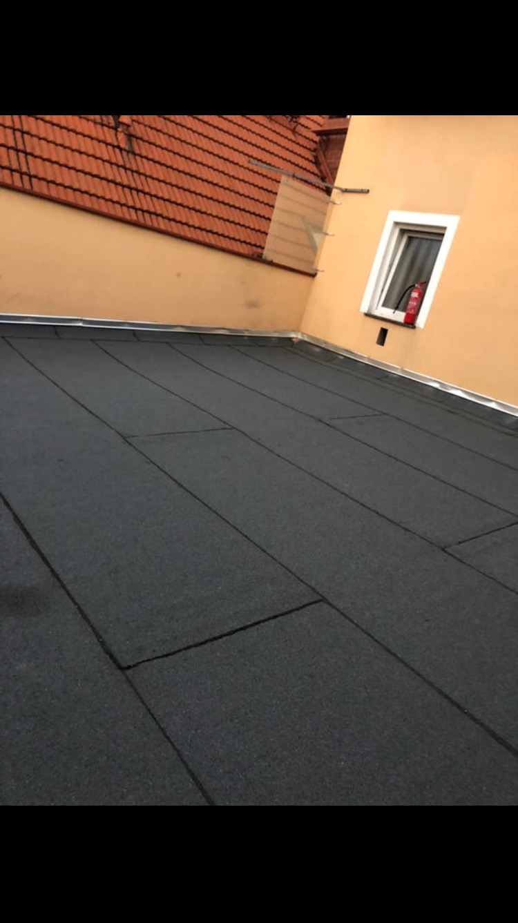 Czarna, płaska powierzchnia dachu z papy bitumicznej, widok z góry, z fragmentem ściany budynku w kolorze kremowym i oknem z gaśnicą w tle oraz fragmentem dachu z czerwonej dachówki ceramicznej.