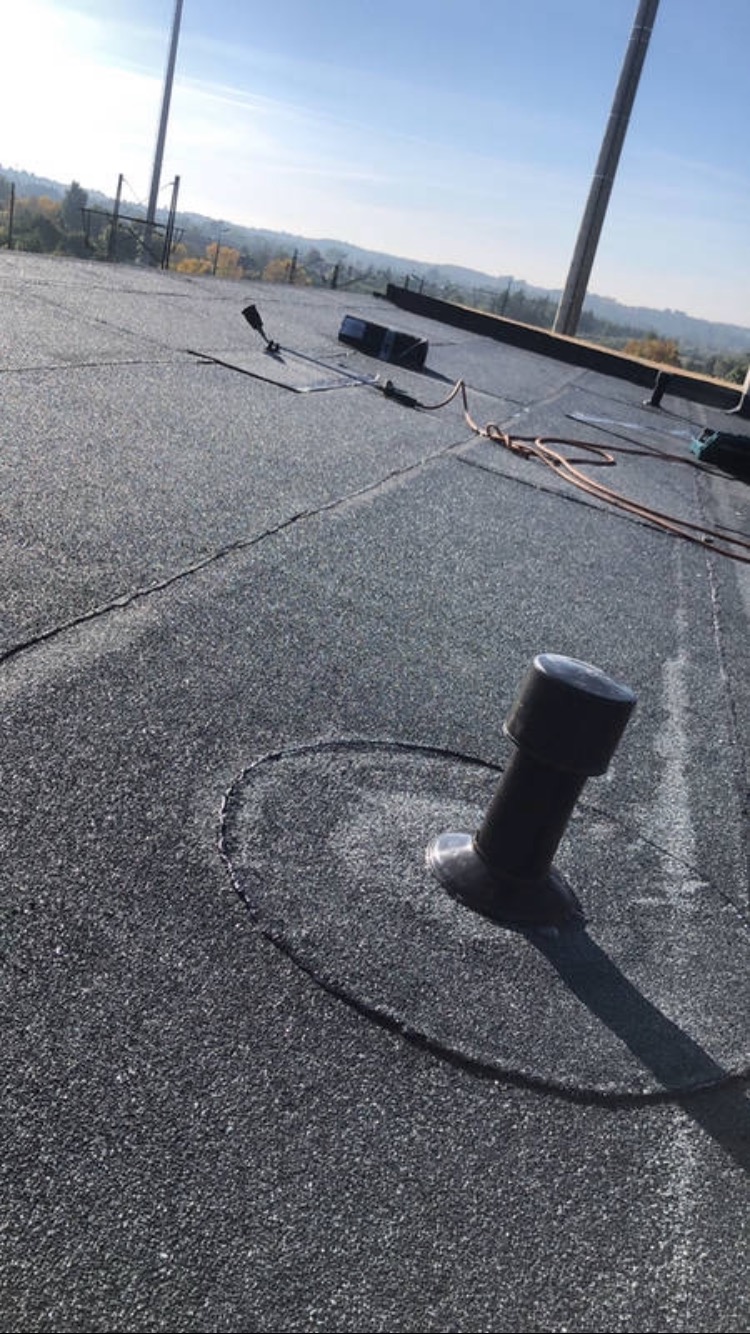 Szary, płaski dach pokryty papą z widocznym kominkiem wentylacyjnym i narzędziami dekarskimi w tle, krajobraz zalesionych wzgórz pod błękitnym niebem.