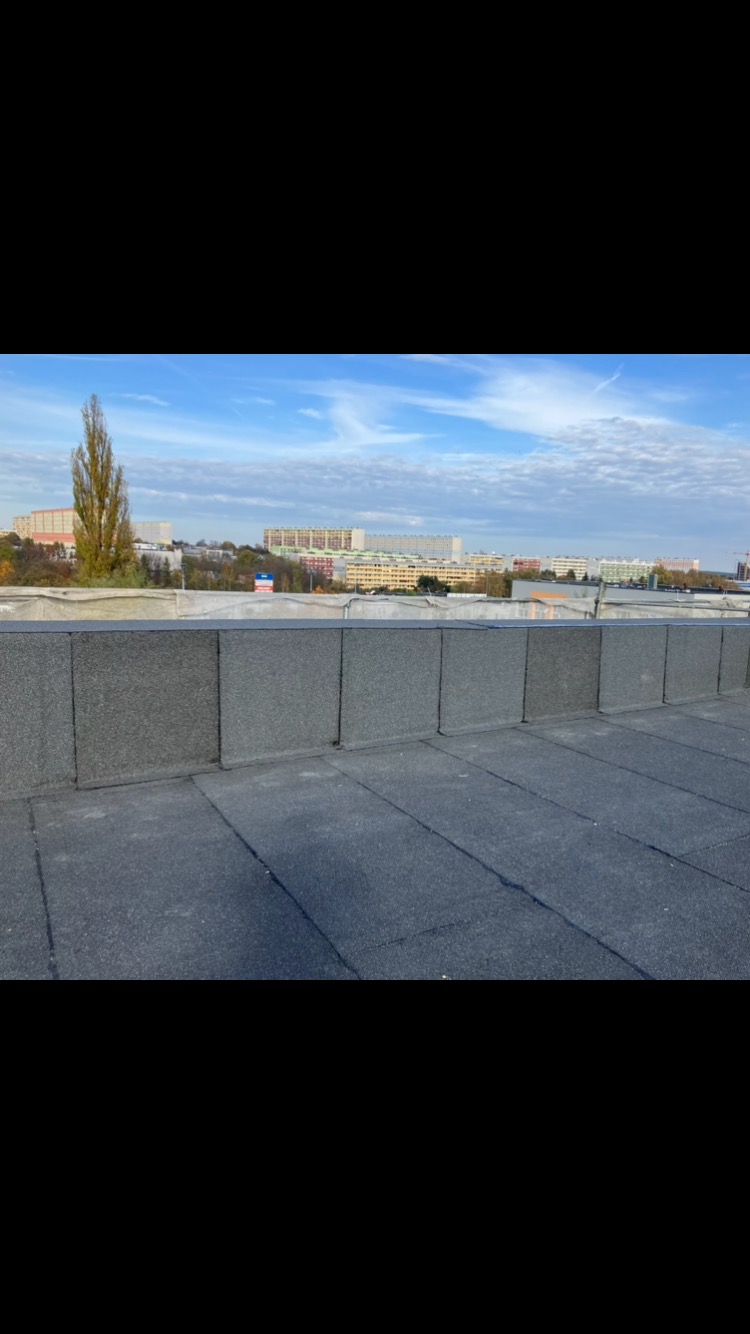 Płaski dach pokryty papą termozgrzewalną z obróbkami blacharskimi na tle miejskiego krajobrazu w słoneczny dzień.