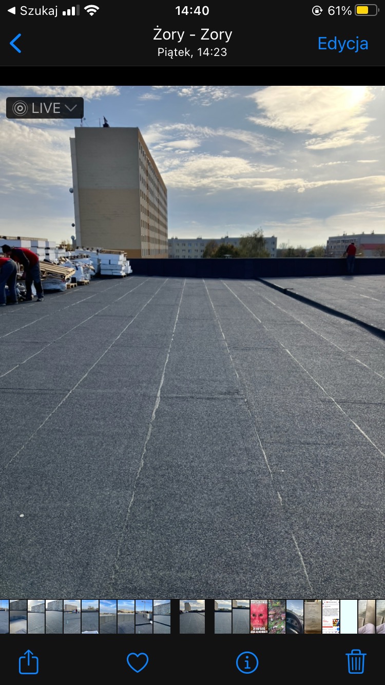 Ekipa dekarzy na płaskim dachu budynku, przygotowuje materiały izolacyjne, widoczny budynek w tle.